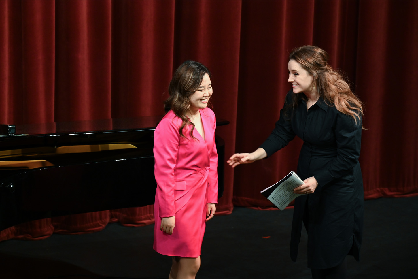 A female student, wearing a bright pink dress, standing in front of a piano, with a women with brown hair and wearing a black dress, smiling at the student, walking on stage.